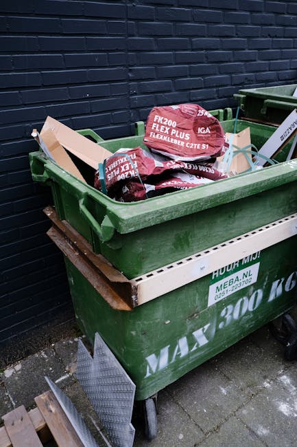 A large green wheeled waste bin filled with bags of construction or renovation debris, including red and white polypropylene bags labeled for insulation or flexible material, placed on a paved area next to a dark brick wall. The bin is stacked over another green and white commercial waste container, which has identification and handling information printed on its side. The scene suggests on-site waste disposal related to renovation or building work, with the company Rubbish Removal Pimlico potentially involved in the collection or management of the debris, illustrating an example of alternative waste handling for builders' waste removal after renovations in an urban setting.