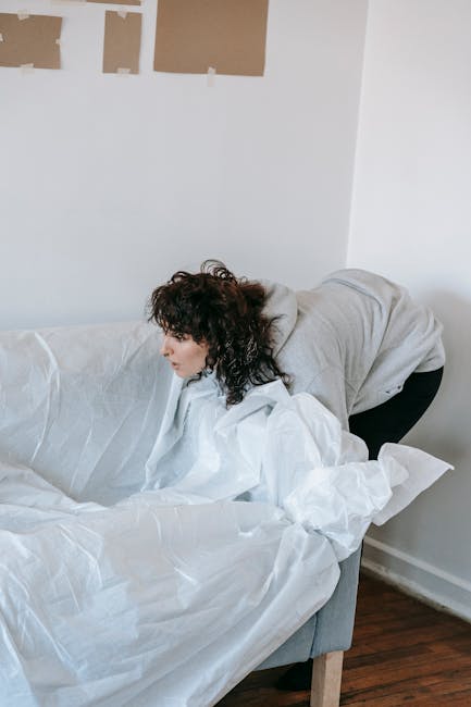A woman with curly dark hair, wearing a light grey hoodie and black trousers, is leaning over a white sofa with a textured fabric cover in a well-lit room. She appears to be in the process of arranging or removing white bedding or cushions from the sofa. The background shows a plain white wall with three rectangular patches of beige wallpaper or paint near the corner. The room has a dark wooden floor, and natural light illuminates the scene, creating soft shadows. The setting suggests a residential interior, possibly during a tidying or cleaning activity, which aligns with the context of rubbish and waste handling services. Rubbish Removal Pimlico could offer relevant solutions for disposing of bedding or soft furnishings when upgrading or clearing space, emphasizing the importance of professional waste management for household clutter or bulky furniture removal.