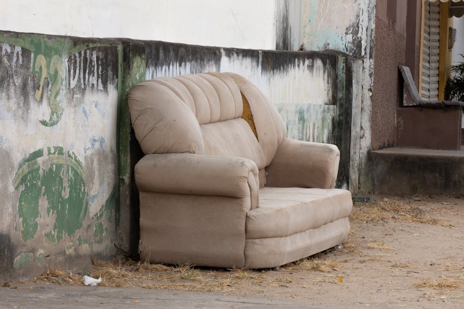 A beige, fabric-upholstered armchair with visible wear and tear, including noticeable damage to the upper backrest where the fabric is torn and yellowish padding is exposed. The armchair is positioned outdoors against a weathered concrete wall, which is painted with faded green and white markings and shows signs of moss, dirt, and peeling paint. The setting appears to be in a residential or urban environment, with a narrow gravelly ground surface scattered with small debris and dry leaves. To the right, there is a small brick step leading up to a partially visible doorway with a closed shutter or door, and some adjacent foliage can be seen. The scene, captured in natural lighting, suggests an instance of private waste handling or on-site clearance involving the removal of furniture, possibly managed by Rubbish Removal Pimlico, for alternative rubbish disposal options outside regular municipal collection.