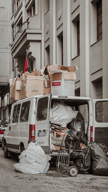 A white panel van parked on a city street during daytime, with its rear doors open revealing a load of discarded household waste and clutter. Inside the van, large black and white garbage bags are piled alongside flattened cardboard boxes, broken wooden pallets, and miscellaneous debris. Some waste is spilled partially onto the pavement, including additional bags leaning against the van’s open doors and scattered on the ground nearby. The van is positioned adjacent to a multi-storey building with a light beige or grey facade, featuring rectangular windows and decorative architectural elements. The scene suggests temporary on-site clearance or private waste collection, handled by Rubbish Removal Pimlico, situated in a dense urban environment with surrounding structures rising alongside. The natural lighting indicates an overcast day, giving a subdued tone to the scene while emphasizing the utilitarian nature of waste removal or rubbish disposal activities.