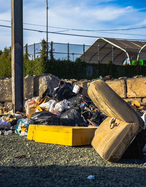 Two large black plastic rubbish bags and a cardboard box containing waste are placed on a sidewalk next to a black metal fence and a gravel driveway, with trees and a building partially visible in the background. The bags appear crumpled and filled with household or garden waste, while the cardboard box is marked with printed text and slightly torn. The scene is outdoors, in an urban environment, illuminated by natural light, and the items are positioned close to the curb, ready for collection or disposal. This scene reflects an example of private waste handling that Rubbish Removal Pimlico provides as part of their rubbish removal services in Pimlico.