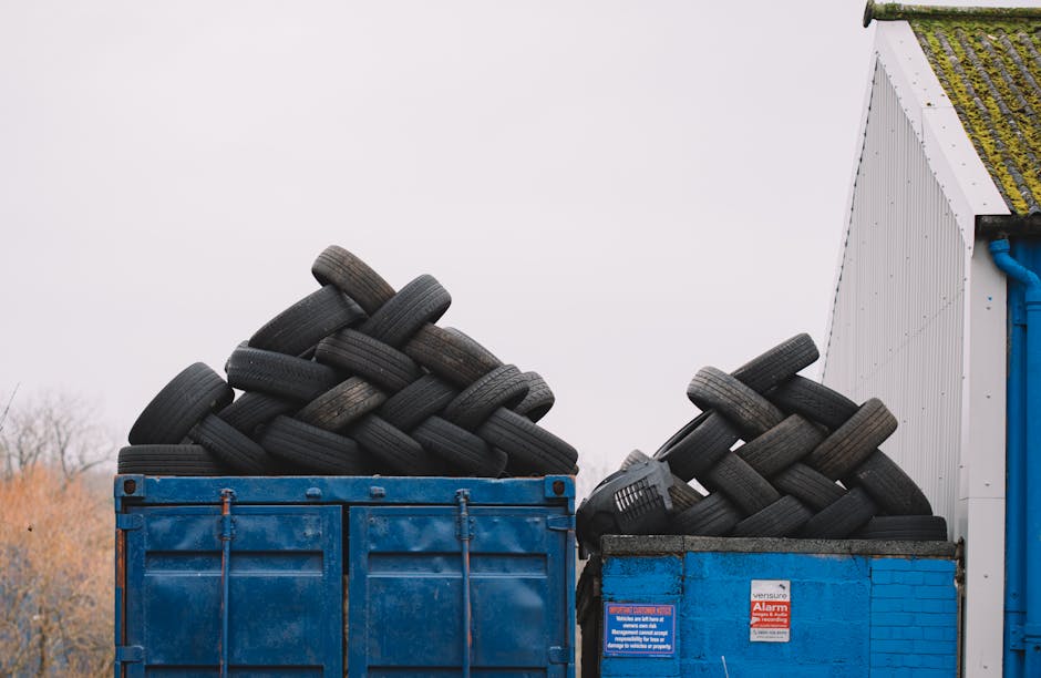 An outdoor scene showing two large open containers filled with used black rubber tires, stacked and arranged in an irregular manner. The left container is a blue metal skip with slightly rusted edges and a weathered surface, containing a significant pile of tires that extend above the container's rim. To the right, a smaller blue brick enclosure also holds a smaller amount of tires, with some tires leaning against the brick wall, positioned near a building with a white, slightly weathered corrugated metal exterior, moss or algae growth visible on the roof edge. In the background, the sky is overcast with a uniform light grey tone, and faint trees or shrubbery are visible in the distance. The scene suggests a private or alternative waste handling or tyre disposal area, relevant to rubbish removal services specializing in large or bulky item collection, as indicated by the company's mention of rubbish in their domain.