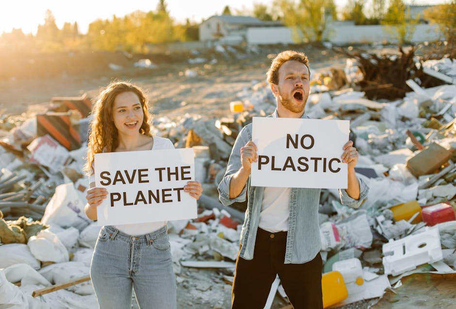 In the foreground, a young woman with wavy, shoulder-length brown hair, wearing a white t-shirt and light blue jeans, holds a sign reading ‘SAVE THE PLANET’ while smiling. Beside her, a young man with short, curly red hair and a beard, dressed in a light denim jacket over a white t-shirt and dark trousers, holds a sign that states ‘NO PLASTIC’ with a serious expression and mouth open as if speaking. They are standing amidst a large pile of discarded plastic waste, including bottles, containers, and packaging materials, situated outdoors on a dirt and gravel surface with scattered debris. In the background, there are leafless trees, a few small buildings, and a partly cloudy sky during late afternoon, with warm sunlight casting a golden hue over the scene. The image implicitly emphasizes issues related to waste management, environmental pollution, and the importance of responsible rubbish disposal, aligning with the topic of alternative waste handling and independent collection services often provided by companies like Rubbish Removal Pimlico.