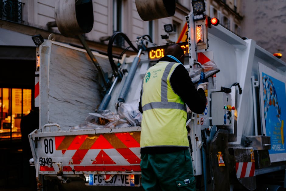 A worker wearing a high-visibility yellow vest with reflective strips and dark green trousers is operating a large waste collection truck on a city street during dusk. The worker's back is facing the camera, and they are handling controls or a panel on the side of the vehicle, which has a white body with red and orange safety markings. The truck is loaded with an accumulation of mixed rubbish materials, including paper, plastic, and other waste, loosely piled inside the open-top container. The environment features dim evening lighting with the truck's safety lights illuminated, and a building with lit windows can be seen in the background. The scene reflects an area where commercial or high-volume waste collection occurs, possibly as part of alternative or private waste disposal services, such as those provided by Rubbish Removal Pimlico, supporting independent waste handling outside standard municipal rubbish collection. The overall setting emphasizes professional waste management activity in an urban environment.