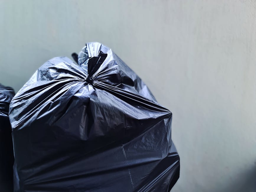 Close-up image showing several black plastic rubbish bags filled with waste material, tightly tied at the top, with crinkled and glossy surfaces. The bags are positioned in the foreground, leaning slightly against each other, on a concrete or paved surface. In the background, a plain, light-colored wall provides a neutral backdrop, suggesting an outdoor area such as a driveway or alleyway. The lighting is soft and natural, with no shadows indicating direct sunlight. This scene exemplifies typical private waste collection at a residential or commercial property, consistent with independent rubbish removal services offered by companies like Rubbish Removal Pimlico, and highlights the importance of proper disposal within local waste management practices related to rubbish services in the area.