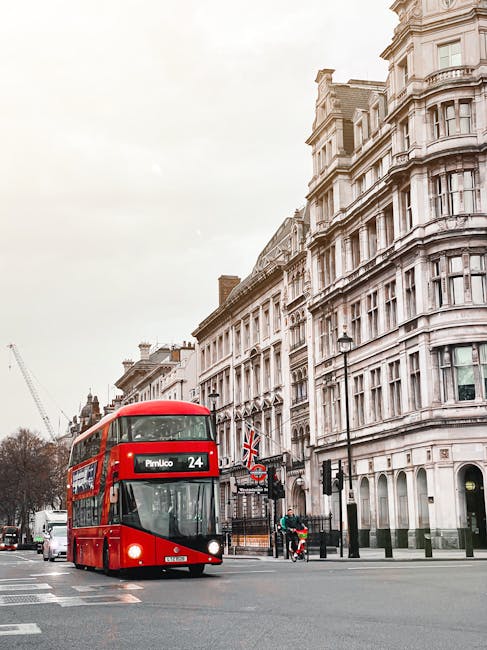 A double-decker red bus traveling along a city street with historic multi-storey buildings featuring intricate architectural details and large bay windows on the right side. The bus displays the route number 24 and destination 'Pimlico' on its front panel. In the background, a crane is partially visible indicating ongoing construction. The street environment includes a black lamppost, traffic lights, and a newspaper stand, with a few pedestrians and cyclists nearby. The scene is set under a pale, overcast sky, emphasizing the contrast between the bright bus and the elegant stone facades of the surrounding buildings, which are characteristic of traditional London architecture. This urban setting reflects the city's mixed historic and modern transport infrastructure, relevant to private and independent rubbish collection services that operate within such tightly packed and characteristic city environments, like those offered by Rubbish Removal Pimlico that support alternative waste handling methods in the area.