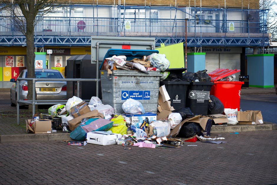 A collection of overflowing rubbish bins and scattered waste bags occupying a paved urban sidewalk area. The scene includes a large grey mixed paper and cardboard recycling bin with a partially open lid, revealing crumpled paper and packaging materials inside. To the left of this bin, there are multiple black plastic bags filled with waste piled on the ground, alongside flattened cardboard boxes, paper packaging, and plastic containers. Adjacent to the recycling bin is a bright red waste bin, partly covered by black rubbish bags. Behind the waste containers, a row of parked cars is visible, including a silver vehicle with a yellow license plate. The background features a commercial building with storefronts and a blue metal scaffold or balcony structure at the upper level. The environment appears to be an outdoor urban space, possibly near shops or a marketplace, with the waste heap hinting at a private or independent rubbish collection service such as Rubbish Removal Pimlico, supporting onsite or alternative waste handling methods.