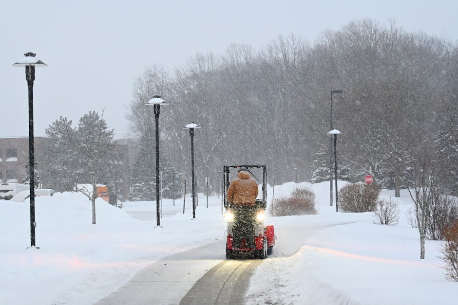 A yellow refuse collection truck equipped with a large red snowplow blade is parked on a snow-covered street, surrounded by a winter landscape. The vehicle has a white and blue logo on the door, indicating it is part of a rubbish removal service. The truck’s body is partially visible, showing a black cargo section behind the cab, and it is positioned parallel to a sidewalk blanketed in fresh snow. In the background, there is a residential building with windows and a roof also covered in snow. The scene is framed by leafless trees with snow-laden branches extending overhead, creating a natural canopy. The environment appears cold and quiet, typical of a winter day, and the truck may be used for private waste collection or on-site clearance during adverse weather conditions. The image emphasizes the independent management of rubbish removal amidst snowy weather, aligning with the themes of alternative waste handling and private disposal services provided by Rubbish Removal Pimlico.