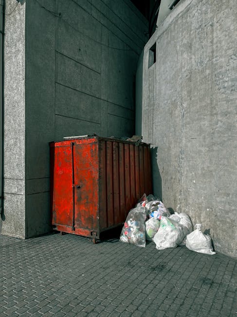 A large, rectangular metal waste container with a rusted, reddish-brown finish sits against the bottom of a dark, smooth wall on a paved urban sidewalk. The container appears weathered, with visible patches of rust and peeling paint. Next to it, several clear plastic bags filled with mixed rubbish, including paper and plastic bottles, are piled on the dark grey cobblestone surface. The background features a textured, light grey concrete wall on the right and a darker, smoother wall on the left, with a small, narrow window or opening visible high above near the top right of the image. The scene is illuminated by natural daylight, casting subtle shadows on the ground and walls, suggesting an outdoor setting designated for rubbish collection or disposal, likely managed by a private waste removal service such as Rubbish Removal Pimlico, to support alternative waste handling methods in an urban environment.