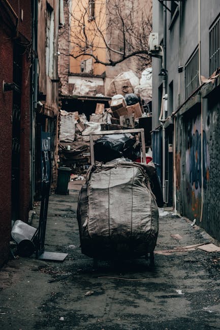 A large, rectangular metal waste container with a rusted, reddish-brown finish sits against the bottom of a dark, smooth wall on a paved urban sidewalk. The container appears weathered, with visible patches of rust and peeling paint. Next to it, several clear plastic bags filled with mixed rubbish, including paper and plastic bottles, are piled on the dark grey cobblestone surface. The background features a textured, light grey concrete wall on the right and a darker, smoother wall on the left, with a small, narrow window or opening visible high above near the top right of the image. The scene is illuminated by natural daylight, casting subtle shadows on the ground and walls, suggesting an outdoor setting designated for rubbish collection or disposal, likely managed by a private waste removal service such as Rubbish Removal Pimlico, to support alternative waste handling methods in an urban environment.