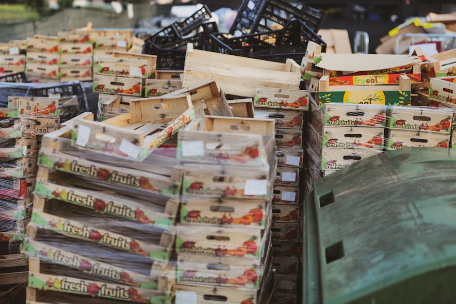 A large outdoor collection area filled with numerous wooden fruit and vegetable crates, predominantly stacked in neat piles. The crates are light-colored, with some bearing colorful printed labels that read 'fresh fruits,' featuring images of various fruits like tomatoes and peppers. In the background, there are additional black plastic crates and some cardboard boxes, indicating a mixture of different waste or recyclable materials. The scene appears to be situated on a paved surface, possibly a yard or loading area, with no visible vehicles. Natural daylight illuminates the scene, casting soft shadows and highlighting the textures of the wooden crates and the various stacking arrangements. This setting could be part of a private waste collection or surplus stock clearance, illustrating a typical scenario where Rubbish Removal Pimlico might handle bulky or non-standard waste items, emphasizing the importance of proper disposal or alternative collection methods for such materials.
